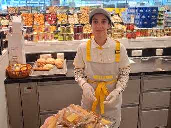 Junge Frau mit Arbeitskleidung von BILLA PLUS steht in einem Suoermarkt in der Feinkostzubereitung. Vor sich hat sie einen Rollwagen mit zubereiteten Weckerln stehen.
